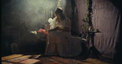 rachel bochner in a dress sitting on the couch, writing in a diary and tearing a page out of it